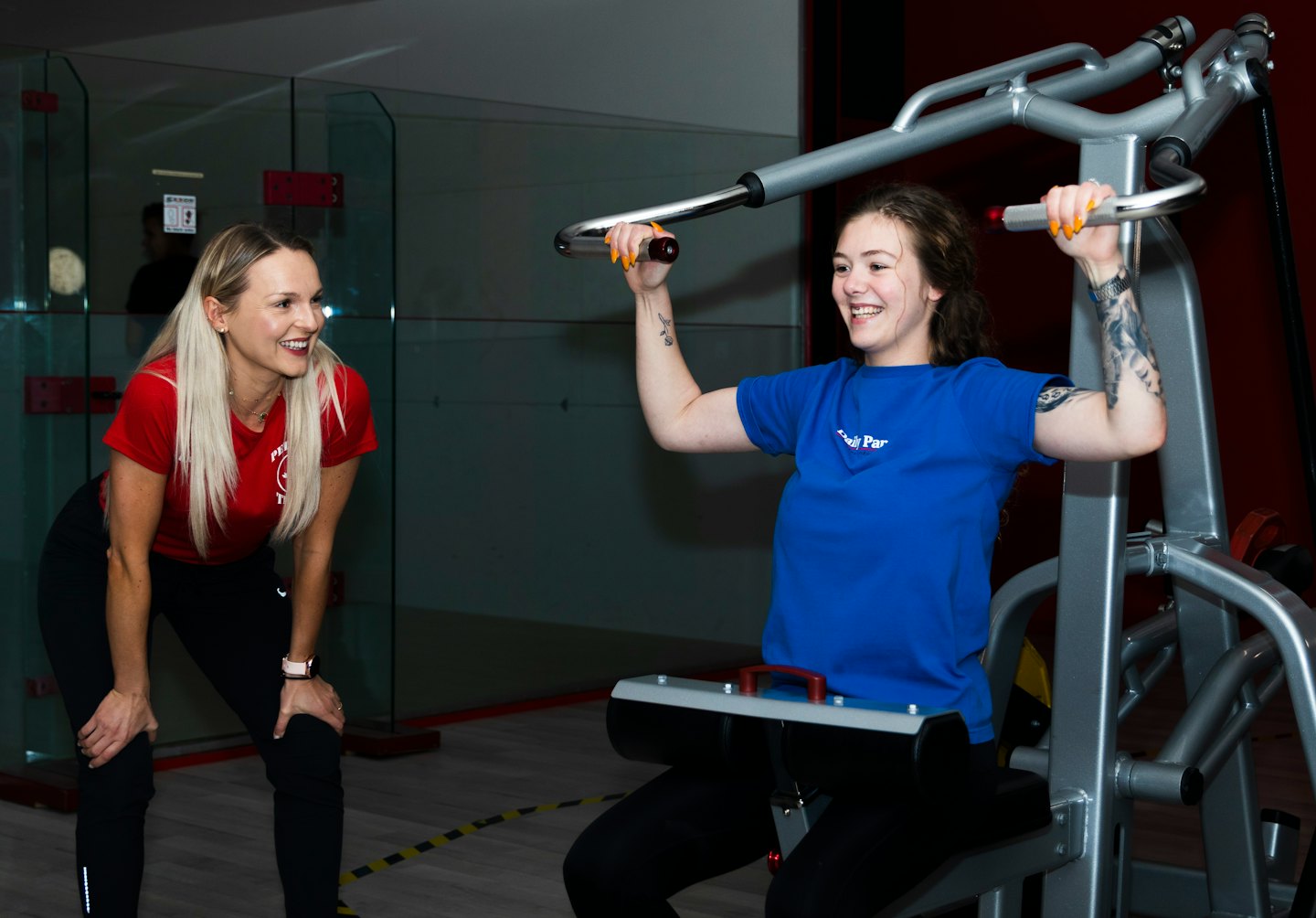 Trainer smiles at client using shoulder press machine.